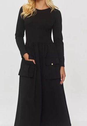 Frau trägt ein schwarzes Langarmkleid mit großen Klapptaschen, eine Hand in einer Tasche, steht vor einem schlichten Hintergrund.