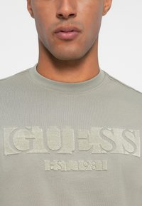 Tricou din bumbac gri, cu un logo texturat "GUESS" în gri deschis. Guler rotund și mâneci scurte, cu detalii de cusătură subtile.