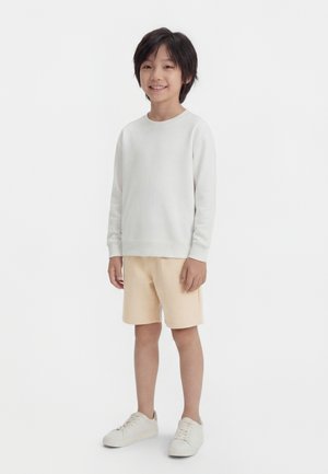 Garçon debout et souriant, portant une chemise blanche à manches longues, un short beige et des baskets blanches, sur un fond blanc.