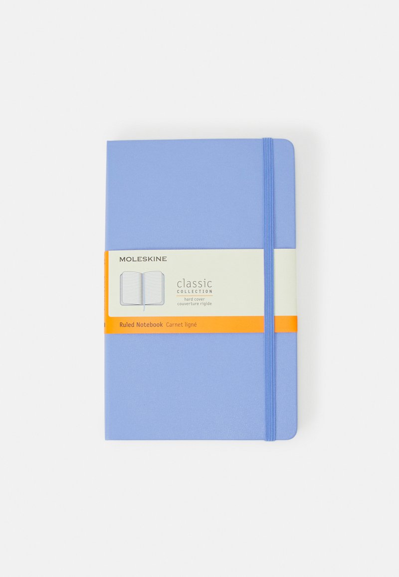 Quaderno Moleskine rilegato in blu chiaro con pagine a righe, tenuto insieme da un elastico blu, con un'etichetta arancione contenente i dettagli del prodotto.