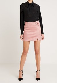 Blouse noire à manches longues avec un nœud papillon, associée à une mini-jupe rose en suédine synthétique avec une fente latérale et un détail d'anneaux en métal argenté.