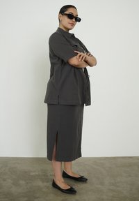 Femme portant une chemise boutonnée gris foncé et une jupe assortie avec une fente arrière, des ballerines noires et des lunettes de soleil, debout les bras croisés.