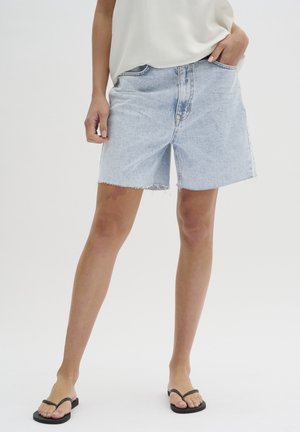 Persoon die lichtblauwe spijkershorts met een ruwe zoom, een wit topje en zwarte teenslippers draagt, staand met één hand in de zak.