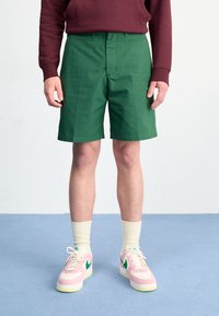 Calções verdes em tecido suave, com frente lisa e bolsos laterais. Combinados com sapatilhas rosas e meias de creme.