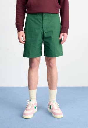 Grønne shorts i et glat stof, med flad front og sidelommer. Båret med lyserøde sneakers og cremefarvede sokker.