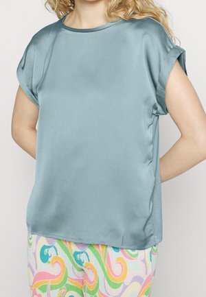 Kvinde iført en lyseblå satinbluse med korte ærmer kombineret med bukser med flerfarvet swirl-mønster mod en ensfarvet baggrund.