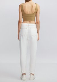 Donna in piedi con la schiena rivolta verso la fotocamera, indossa un top corto beige, jeans bianchi a vita alta e sneakers bianche su uno sfondo neutro.