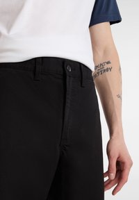 Pantaloni di cotone neri con chiusura a bottone, dal tessuto morbido e dalla vestibilità classica. Un tatuaggio è visibile sul braccio che li tiene.