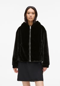 Black faux fur hooded jacket with a zip front, side pockets, and ribbed cuffs; soft texture and relaxed fit. Worn over a black dress.