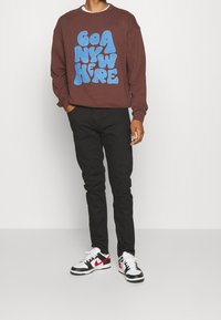 Felpa marrone con grande testo grafico blu "GO ANYWHERE," abbinata a pantaloni neri e sneakers in bianco e nero.
