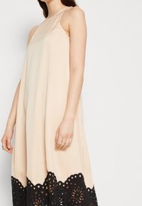Robe sans manches beige avec un col rond, ornée d'une bordure en dentelle noire avec des motifs floraux découpés le long du bas.