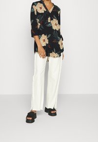 Svart blommig blus med beige blommor och gröna blad, med V-ringning och trekvartsärmar, tillsammans med vita byxor med fransar och svarta plattformssandaler.