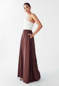 Vrouw in een witte top met één schouder en bruine hoge taille, wijde broek met zakken, staande tegen een effen lichte achtergrond.