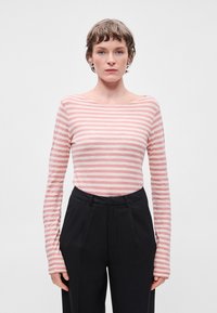 Camisola de mangas compridas às riscas em rosa claro e creme, com um amplo decote à barco. Combinada com calças pretas de cintura alta. Textura suave.