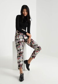Sort langærmet top kombineret med blomster mønstrede, tætsiddende bukser med livlige farver på sort baggrund. Modellen har sorte slip-on sko på.