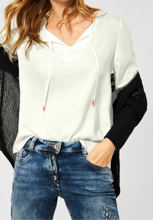 Femme portant un haut à capuche blanc avec cordons, un jean bleu à boutons avec broderie, et un pull en tricot noir drapé sur les épaules.