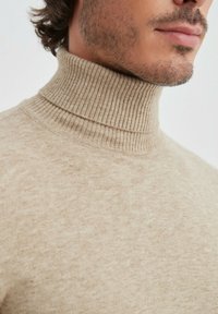 Pull à col roulé beige en tissu doux et texturé avec un col côtelé. Vue en gros plan axée sur l'encolure et le haut du torse.