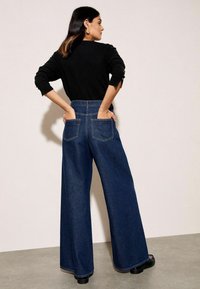 Visoke traperice širokih nogavica u tamnoplavoj boji. Imaju dva stražnja džepa i glatku završnu obradu. Uparene s crnim topom.