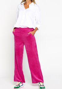 Pantaloni larghi in velluto a coste rosa con tasche frontali, finitura testurizzata, abbinati a una camicia bianca abbottonata e sneakers colorate.