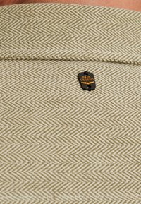 Tissu beige à motif chevrons avec une texture subtile, présentant une étiquette noire avec "PME Legend" en lettres orange attachée.
