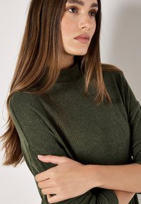 Pull en maille côtelée vert foncé avec col haut et manches longues, mettant en valeur les détails de texture. Les bras du modèle sont croisés dans une pose neutre.