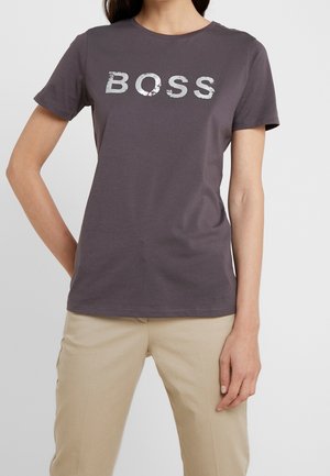 Femme portant un T-shirt gris foncé avec un texte blanc usé "BOSS", associé à un pantalon beige, debout avec une main sur la hanche.
