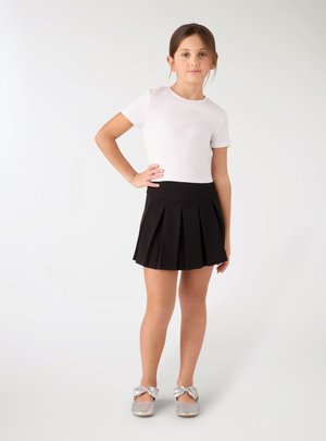 Ragazza giovane in piedi con la mano sul fianco, indossa una maglietta bianca a maniche corte, gonna nera plissettata e scarpe glitter argento con fiocchi.