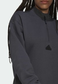 Halvdraget sweatshirt i mörkgrått, tillverkat av mjukt material. Har en hög krage och svart Adidas-logotypdetalj på bröstet.