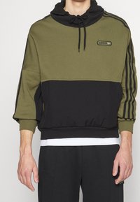 Olivgrön och svart tvåtonspullover med huva, ribbade ärmslut, elastisk nederkant och Adidas-logotyp; har tre svarta ränder på ärmarna.