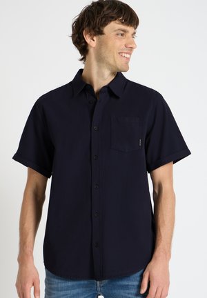 Jeune homme souriant, portant une chemise bleu marine foncé à manches courtes avec une poche poitrine et un jean bleu, tourné vers la droite.