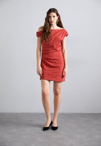 Abito rosso a spalle scoperte, aderente, al ginocchio, realizzato in tessuto liscio. Abbinato a scarpe nere con tacco alto, dal design minimalista.
