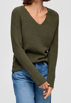 Femme portant un pull en tricot vert olive à col en V avec manches longues et un jean bleu en denim, mains délicatement jointes devant.