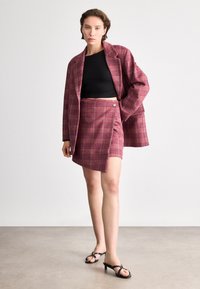 Sarkans skotu raksta žakets un saskaņota svārki, abi ar teksturētu audumu. Melnā piegulošā blūze zemāk un melnas atvērtā purniņa augstpapēžu kurpes.