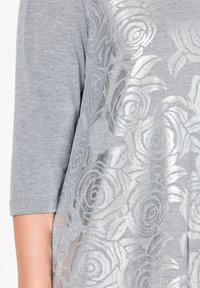 Top grigio con maniche tre quarti caratterizzato da un motivo floreale argentato. Il tessuto presenta una texture liscia con un design metallico riflettente.