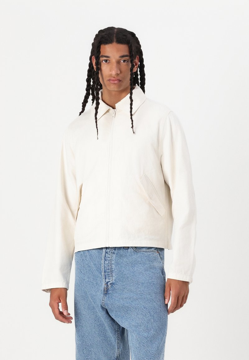 Jachetă albă, ușor texturată, cu fermoar, guler și buzunar frontal, purtată cu blugi denim de culoare bleu deschis. Croială casual, detalii minime.