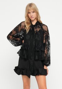 Sort blonderet bluse med peplumdesign, lange gennemsigtige ærmer og sløjfe ved halsen. Sat sammen med en volangsort nederdel.