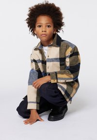 Camicia a quadri in beige, blu e nero con taschino sul petto, abbinata a pantaloni in denim scuro e scarpe nere. Tessuto morbido e vestibilità oversize.