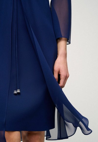 Navy blue sheer dress with long sheer sleeves, featuring a split front design and drawstrings with metallic accents at the hem.