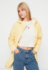 Νεαρή γυναίκα με κίτρινο αδιάβροχο με κουκούλα, λευκό κοντομάνικο πουλόβερ και μπλε τζιν, με τα χέρια στις τσέπες, κοιτάζοντας προς τα αριστερά της, σε λευκό φόντο.