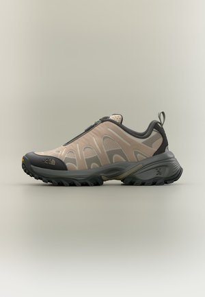 Beiger und grauer Trailrunning-Schuh mit Reißverschluss, robuster Sohle, Mesh-Obermaterial und Zuglasche an der Ferse.