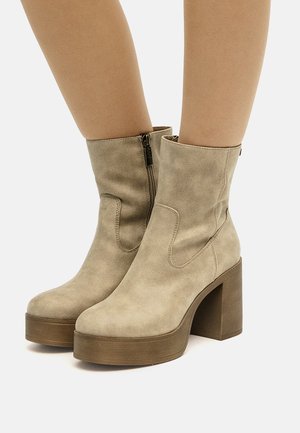 Beige ruskind ankelstøvler med blokhæl, platformsåler og sidelommer glidelåse, båret på bare ben mod en hvid baggrund.
