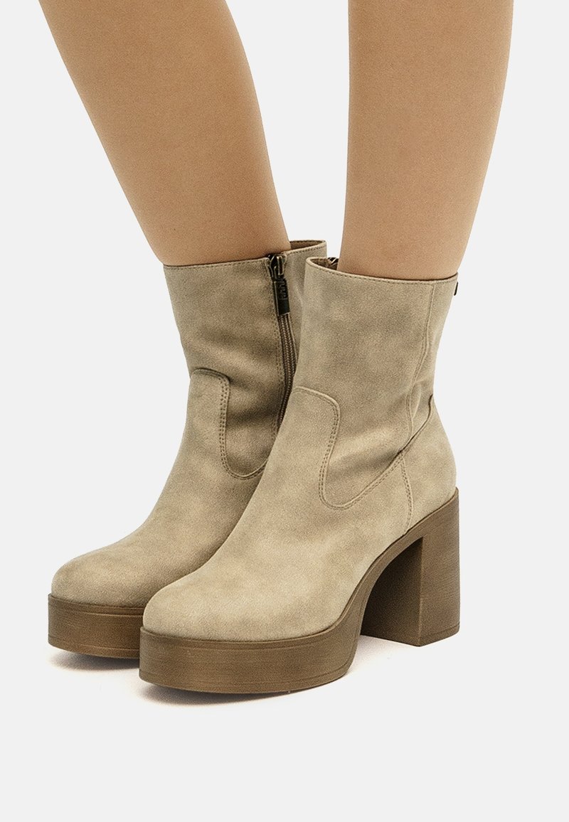 Bottines en daim beige avec talons blocs, semelles plateformes et zips latéraux portées sur jambes nues sur fond blanc.