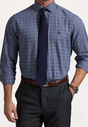 Homme portant une chemise bleue à carreaux avec une cravate bleu marine, une ceinture en cuir marron, un pantalon gris, une montre-bracelet et les manches retroussées jusqu'aux avant-bras.
