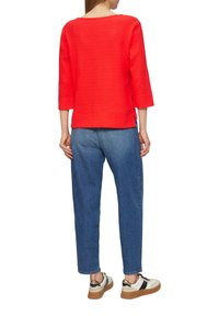Maglia rossa con texture e maniche a tre quarti, abbinata a pantaloni in denim azzurro chiaro e sneaker bianche con dettagli neri e dorati.