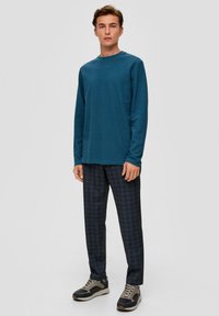 Teal lange mouwen crewneck shirt, gecombineerd met donker geruite broek en atletische sneakers. De stof lijkt zacht; de broek vertoont een gestructureerd patroon.