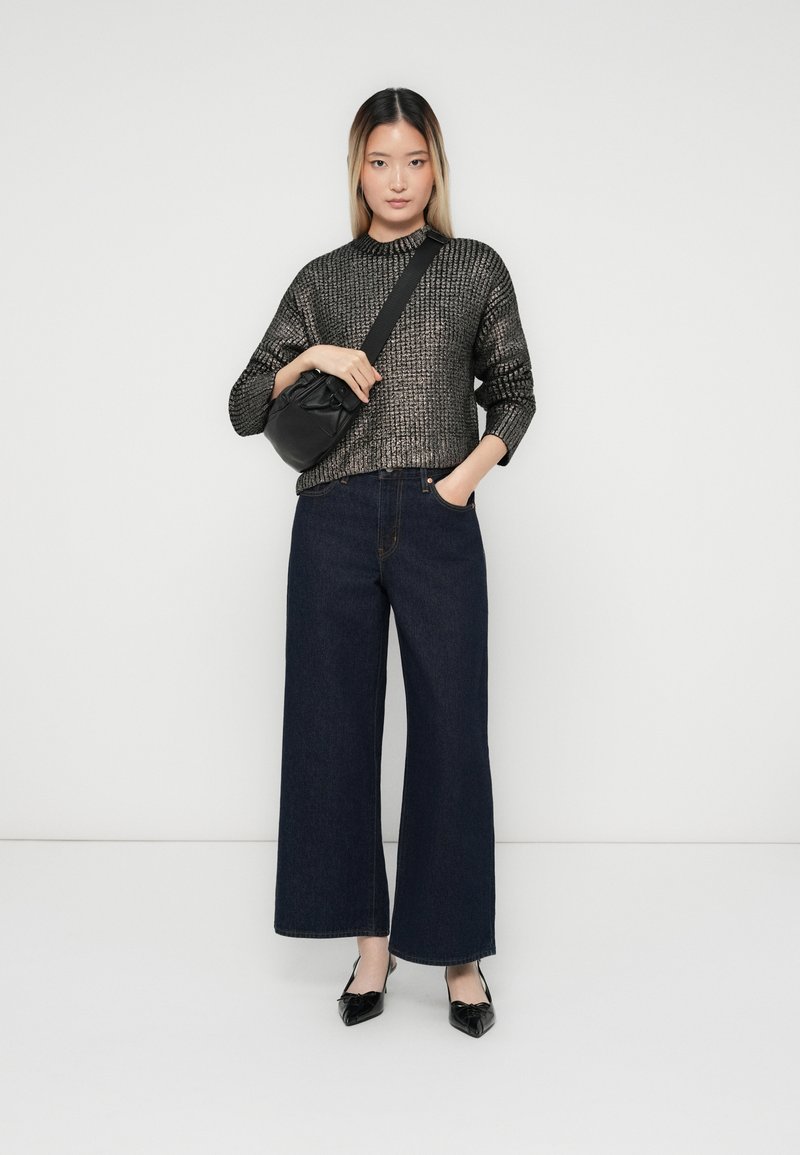 Una donna indossa un maglione in tessuto metallico, jeans scuri a gamba larga, tacchi slingback neri e una borsa a tracolla nera, in un contesto con sfondo bianco.