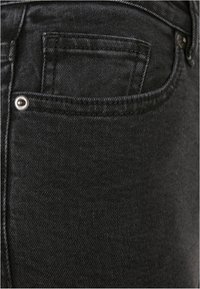 Jeans de mezclilla negros con una superficie texturizada, que presentan un pequeño bolsillo trasero y un detalle de botón plateado. Costuras sutiles visibles.
