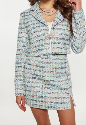 Frau trägt eine pastellblaue, kurz geschnittene Tweed-Jacke und passenden Rock mit kleinem vorderen Schlitz, ergänzt durch geschichtete Halsketten und Ringe.