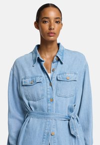 Jasnoniebieska dżinsowa kurtka koszulowa z kołnierzykiem, dwiema kieszeniami na piersi, z przodu zapinana na guziki i z materiałowym paskiem przewiązanym w talii.
