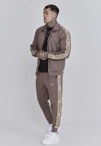 Chaqueta y pantalones de chándal marrones hechos de un tejido suave, con acentos de logotipo beige a lo largo de las mangas y las piernas. Zapatillas blancas completan el look.
